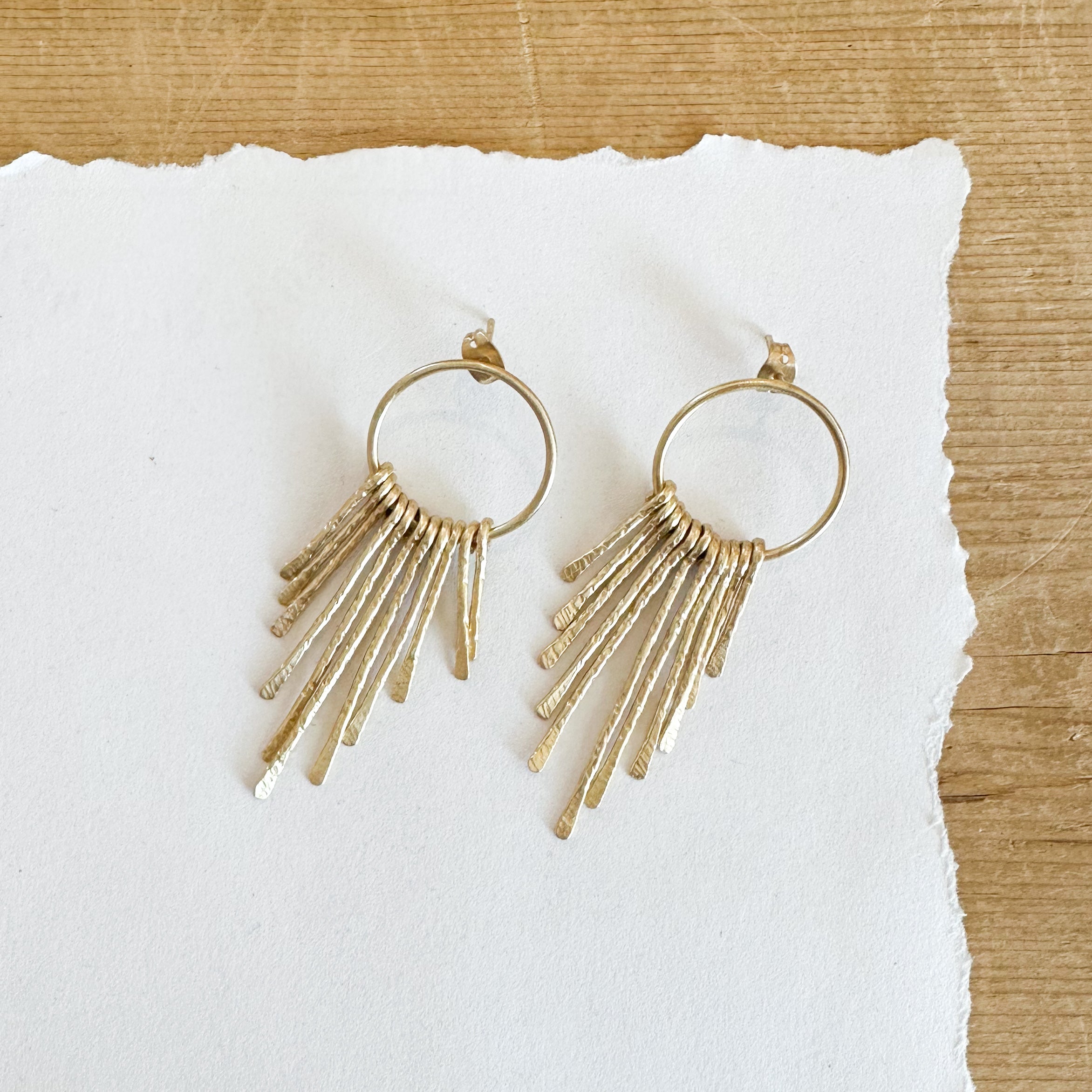 Gold toned earrings featuring hammered pieces that dangle from the hoop forming a fringe on the bottom of the hoop.