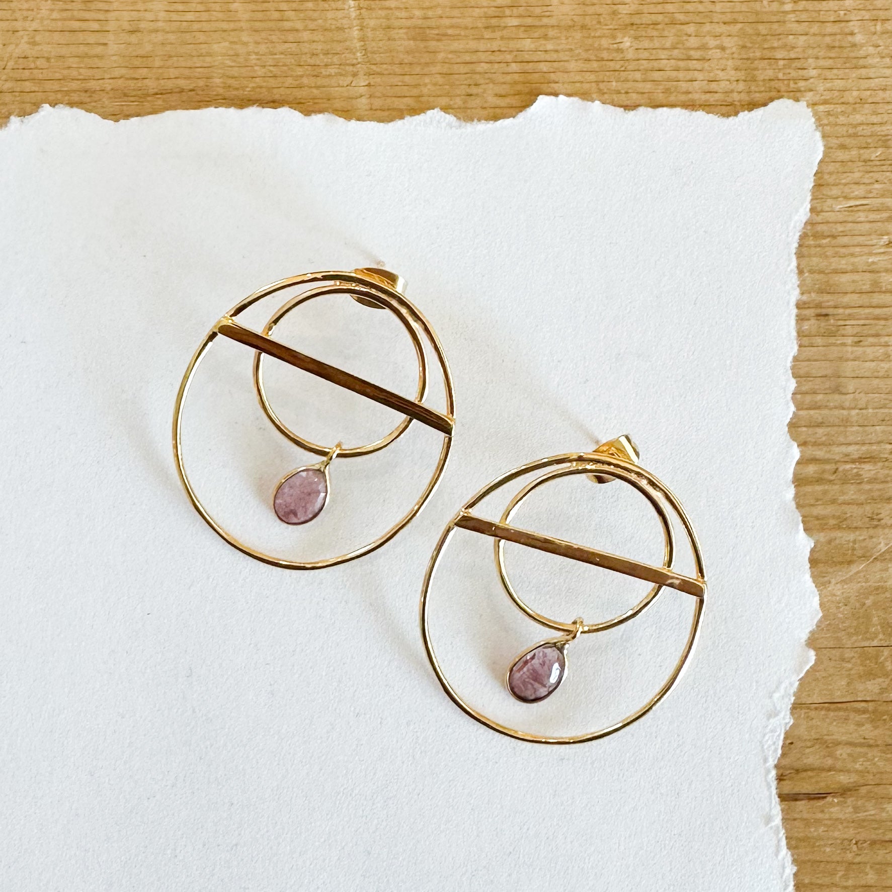 Gold tone double hoop studs with purple toned tourmaline hanging from inner hoop.