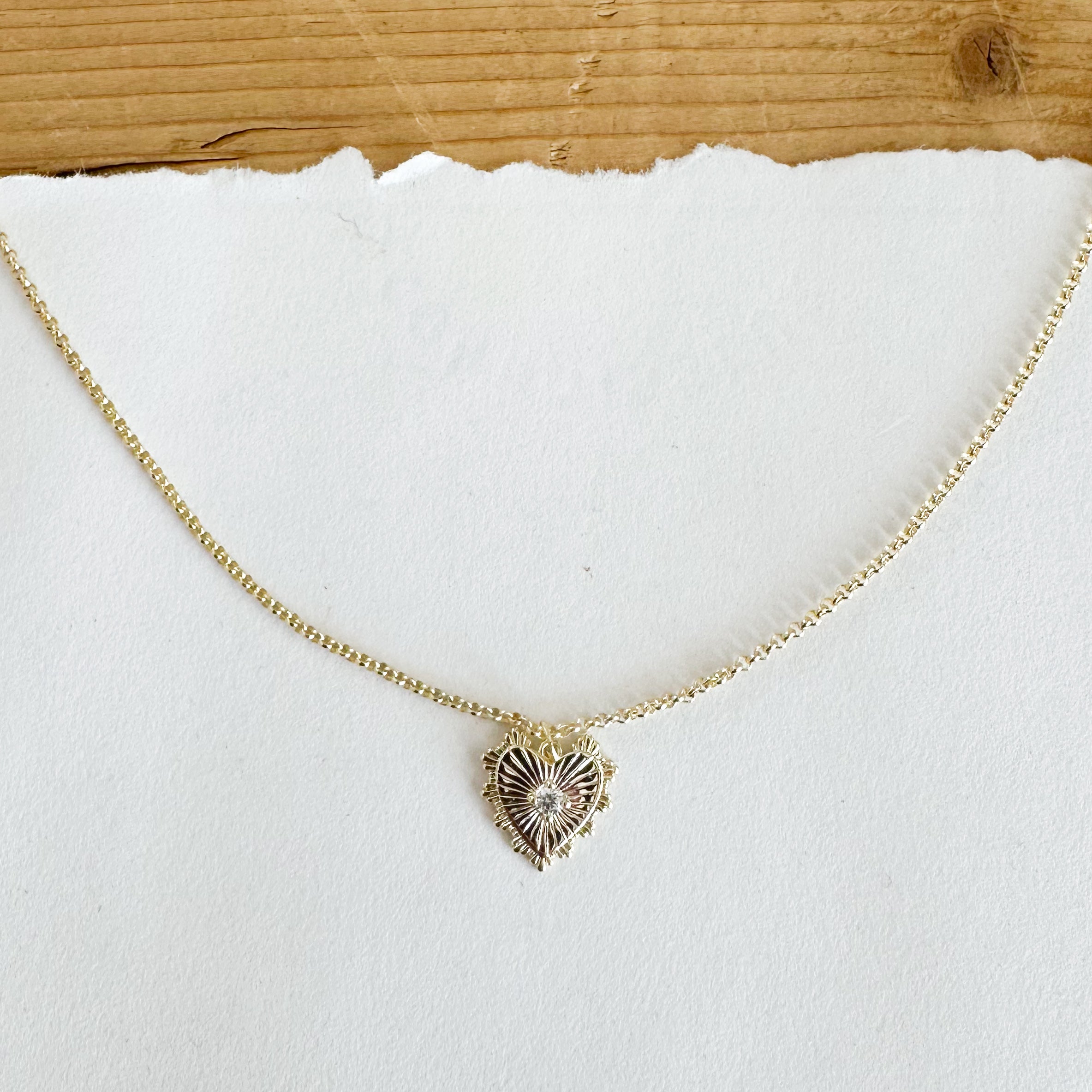 Gold tone necklace featuring a heart charm.  The heart charm is gold toned and features a scalloped edge and a clear round gemstone in the center.